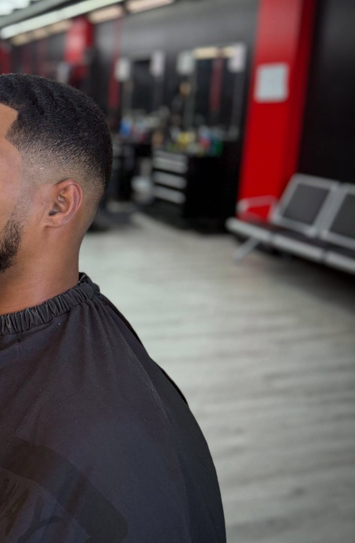 A fresh low fade seen from behind, client in a black cape, red pillars and the waiting bench out of focus